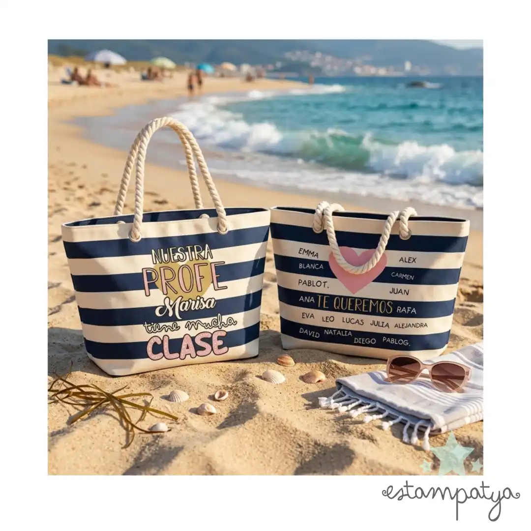 Bolsa Profe tiene mucha clase - Estampatya bolsos de playa de rayas marineras azules y blancas sobre la arena con el mar de fondo. El bolso izquierdo tiene el texto "Nuestra PROFE Marisa tiene mucha CLASE" y el derecho tiene nombres de alumnos alrededor de un corazón rosa con la frase "TE QUEREMOS".