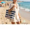 Bolsa Profe tiene mucha clase - Estampatya Una mujer sonriente en la playa lleva colgada al hombro la bolsa de rayas marineras para profesora con el texto "Nuestra PROFE Marisa tiene mucha CLASE". Viste un vestido playero claro, gafas de sol y lleva un libro en la mano, con el mar y la arena de fondo.