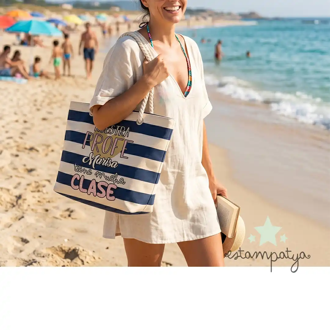 Bolsa Profe tiene mucha clase - Estampatya Una mujer sonriente en la playa lleva colgada al hombro la bolsa de rayas marineras para profesora con el texto "Nuestra PROFE Marisa tiene mucha CLASE". Viste un vestido playero claro, gafas de sol y lleva un libro en la mano, con el mar y la arena de fondo.