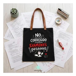 Bolsa de tela negra para profesora con frase divertida sobre corregir examenes y asas marrones, estirada sobre mesa de examenes
